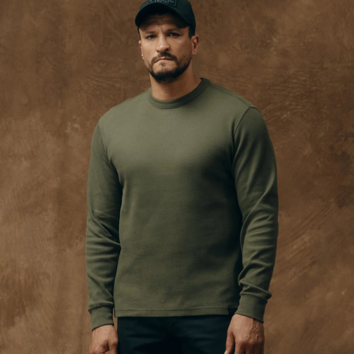 Man wearing a green long-sleeve shirt and dark cap against a brown background