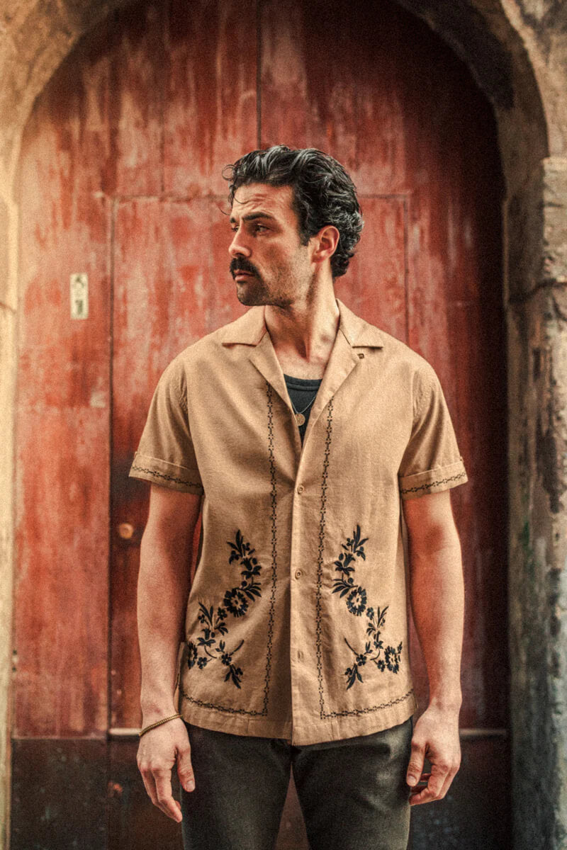 Man wearing a beige shirt with floral embroidery standing in front of a red door.