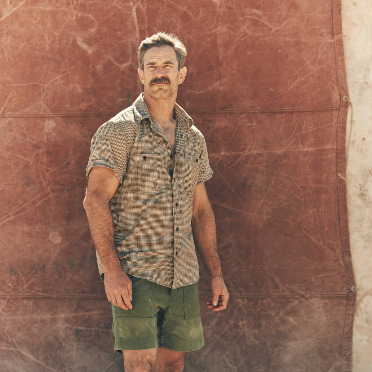 Man wearing a checkered Filson shirt and green shorts standing against a textured brown wall.