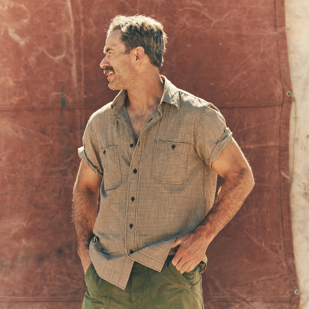 Man wearing a checkered Filson shirt and green pants against a textured wall.