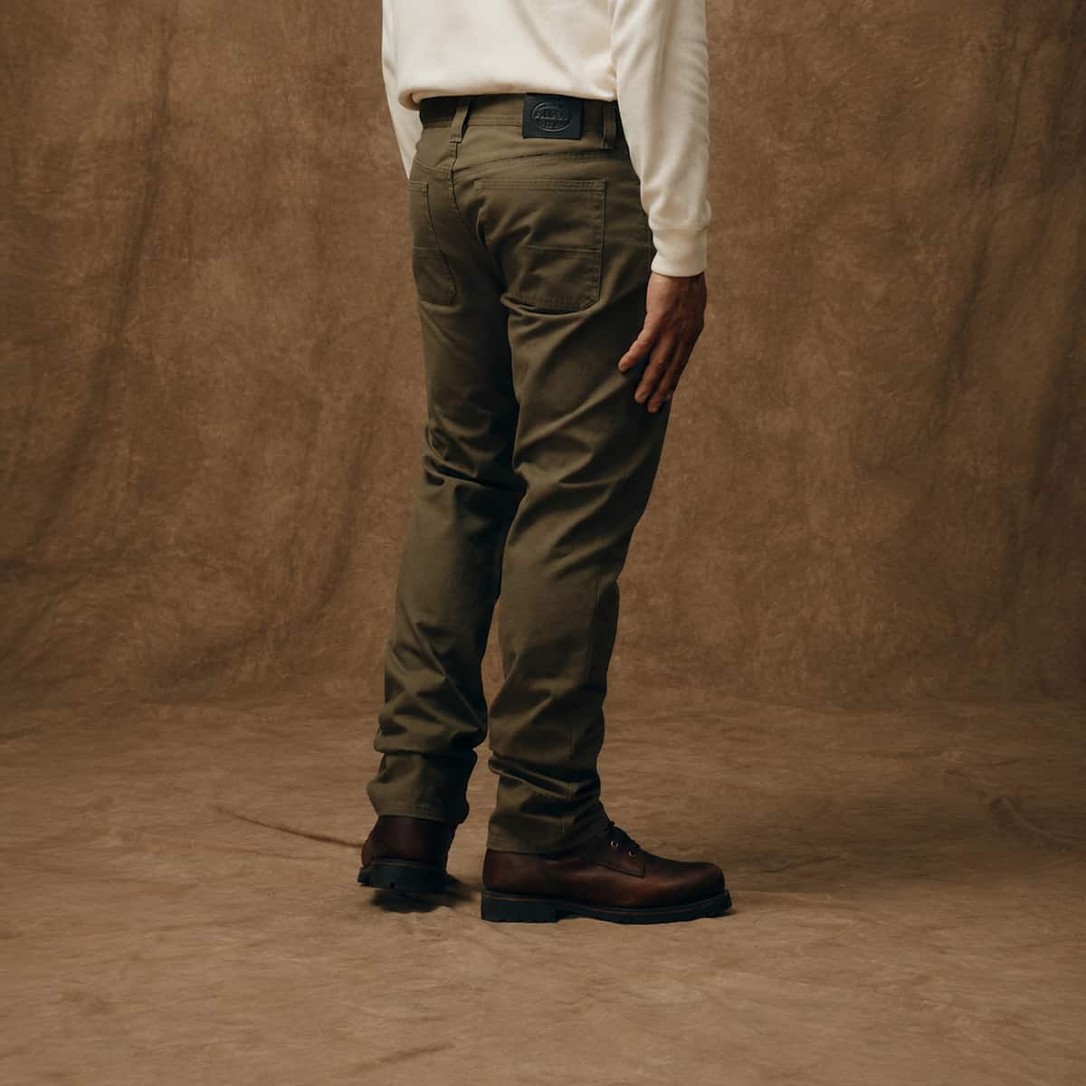 A person shown from the back wearing olive green Filson work pants, a white long-sleeve shirt, and brown leather boots, standing against a textured brown backdrop.
