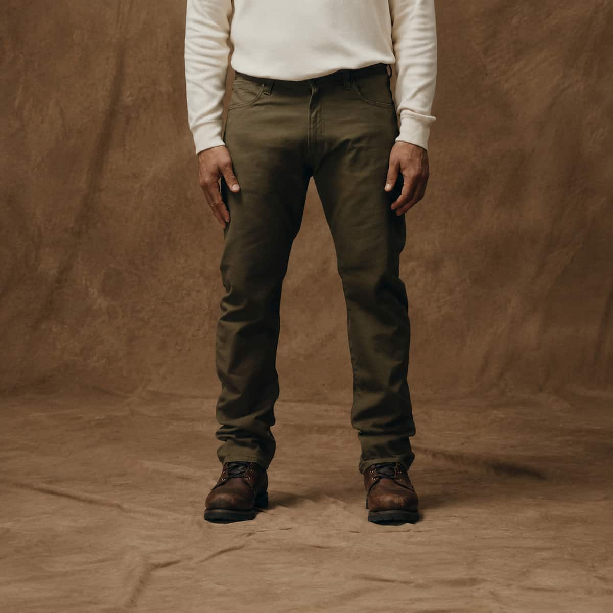 A person wearing olive green Filson 5_Pocket work pants, a cream long-sleeve shirt, and brown work boots, standing against a textured brown fabric backdrop.