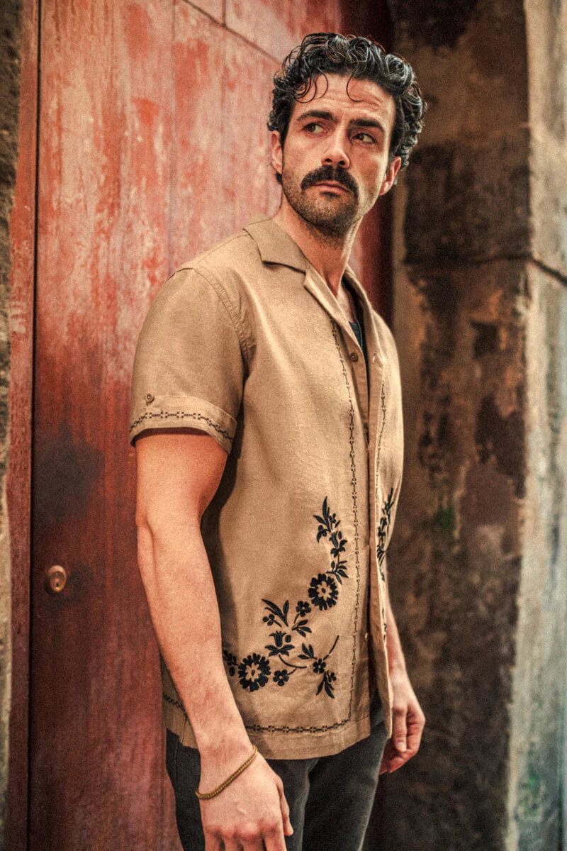 Man wearing a beige shirt with floral embroidery standing against a textured wall.