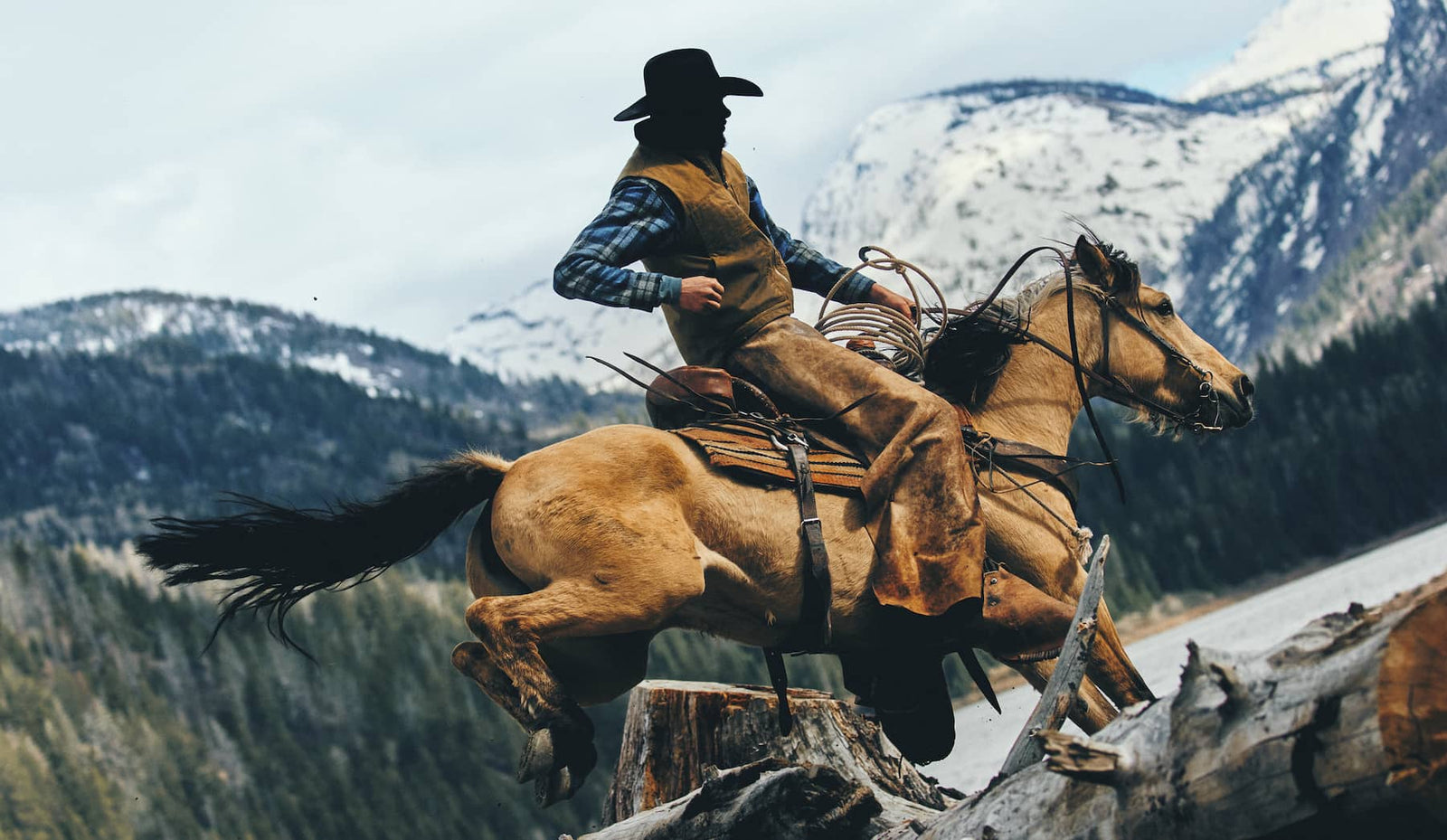 A cowboy in a black hat and brown vest rides a brown horse over a fallen log in a mountain landscape with snowy peaks in the background.