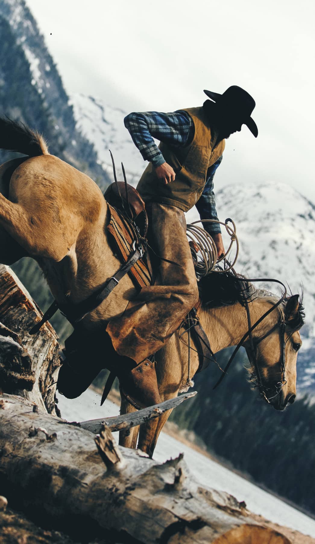 A cowboy in a black hat and brown vest rides a brown horse through a snowy mountain landscape, with another horse and a large fallen log in the foreground.