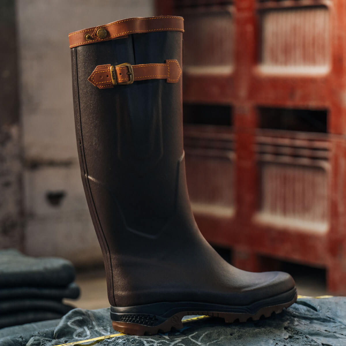 Dark rubber boot with brown leather strap on a textured surface