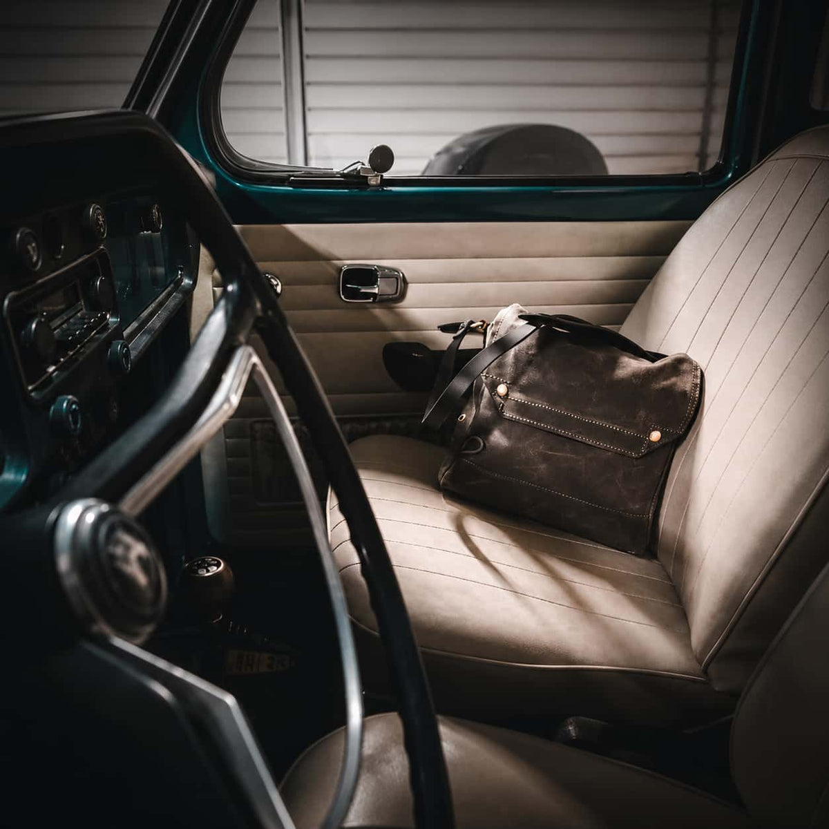 Frost River Archaeologist Satchel with gold-tone hardware rests on the beige ribbed passenger seat of a vintage car interior.