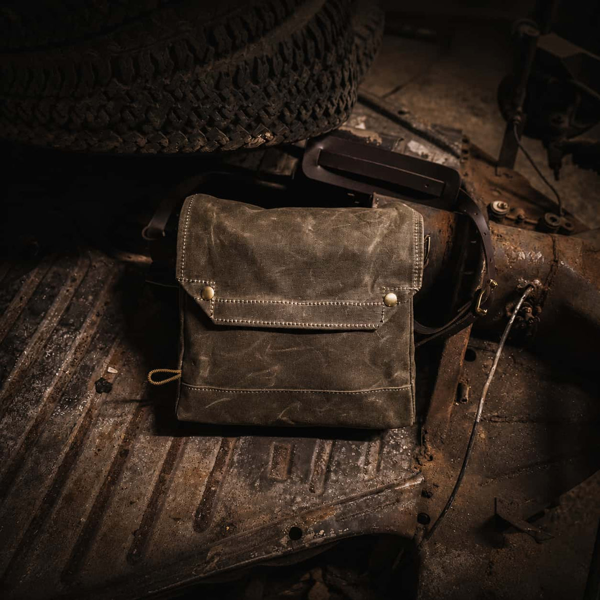 A distressed Frost River Archaeologist Satchel with brass hardware, resting on a rusty metal surface next to a black leather strap.