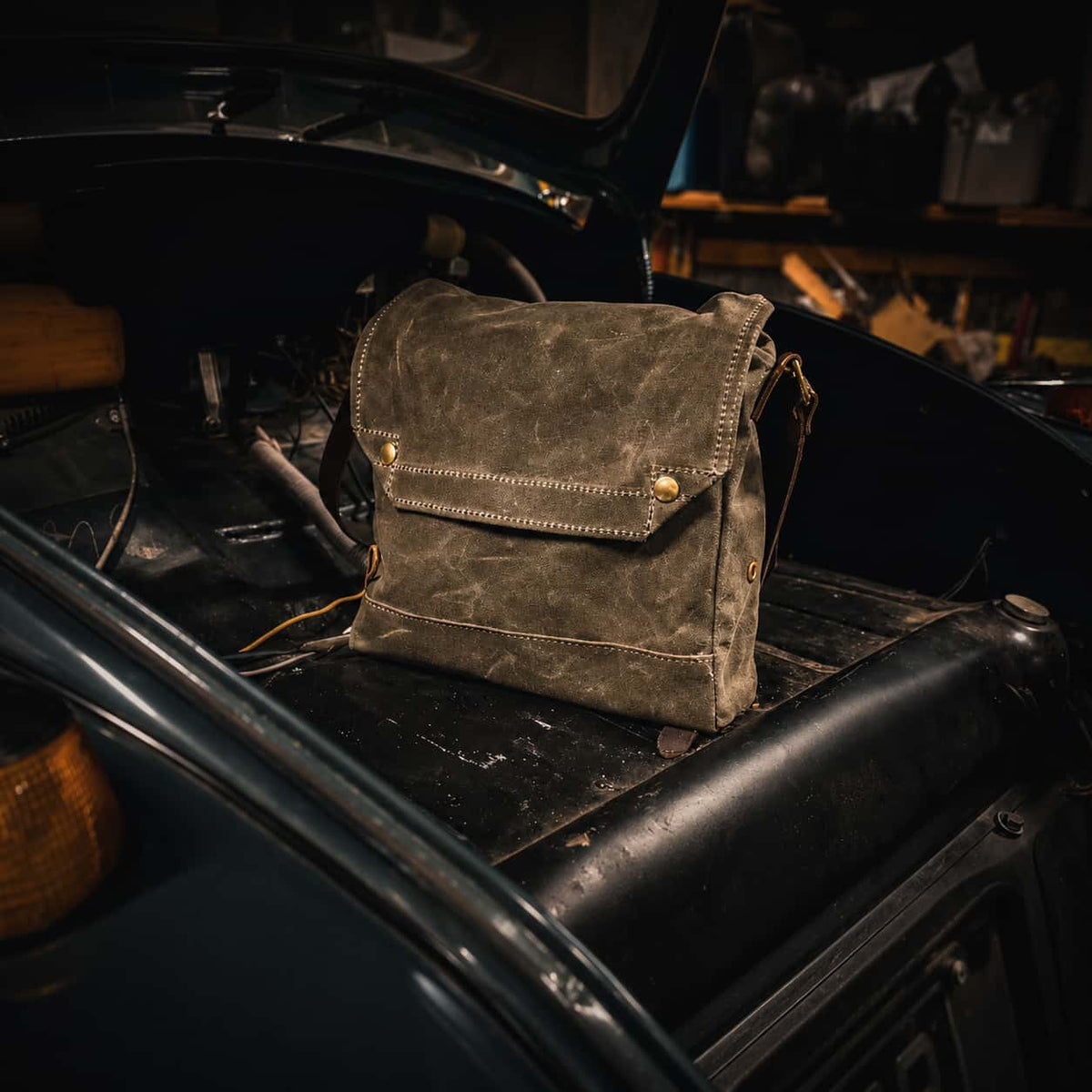 A Frost River Archaeologist Satchel with brass hardware, resting on the open trunk of a vintage black car in a workshop setting.