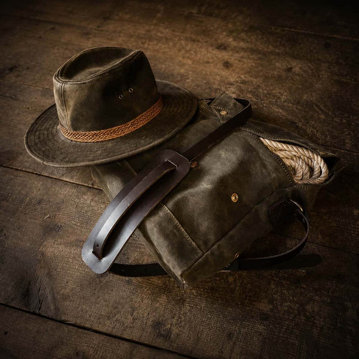A Frost River Archaeologist Satchel with a leather strap and a matching wide-brim fedora hat with a braided band, arranged on a rustic dark wood surface.