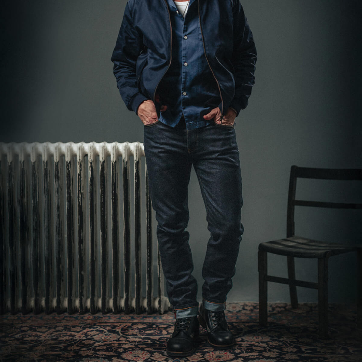 Person wearing a blue jacket and jeans standing in a room with a radiator and chair.