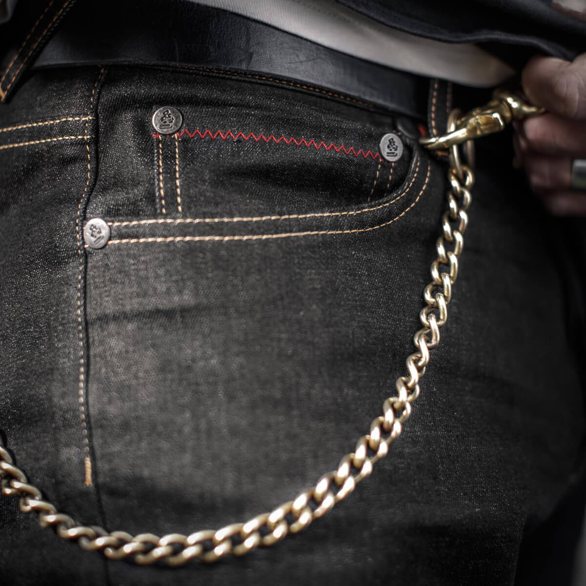 Gold chain hanging from a pocket of dark jeans with red stitching.