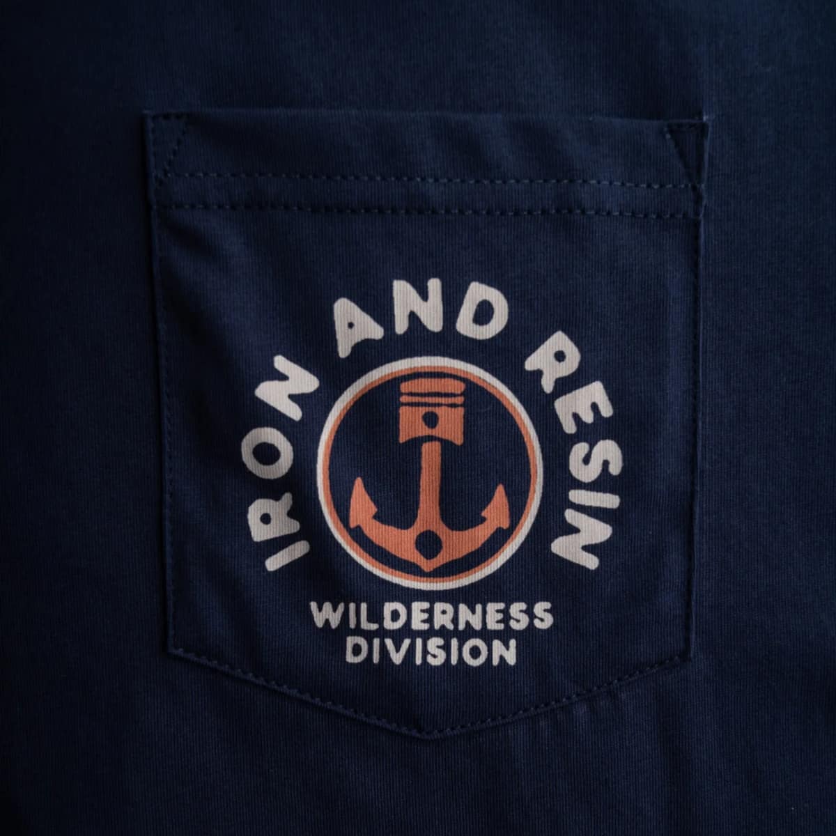 Close-up of a dark navy blue pocket on a matching shirt, featuring a circular embroidered logo with an anchor and the text 'IRON AND RESIN WILDERNESS DIVISION'.