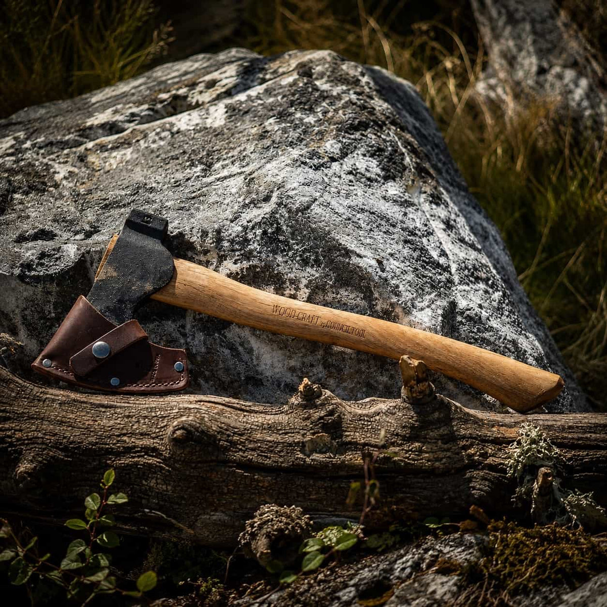 A Camp Carver axe with a light wood handle and dark metal head rests on a log against a large rock outdoors. A brown leather sheath is attached near the head.