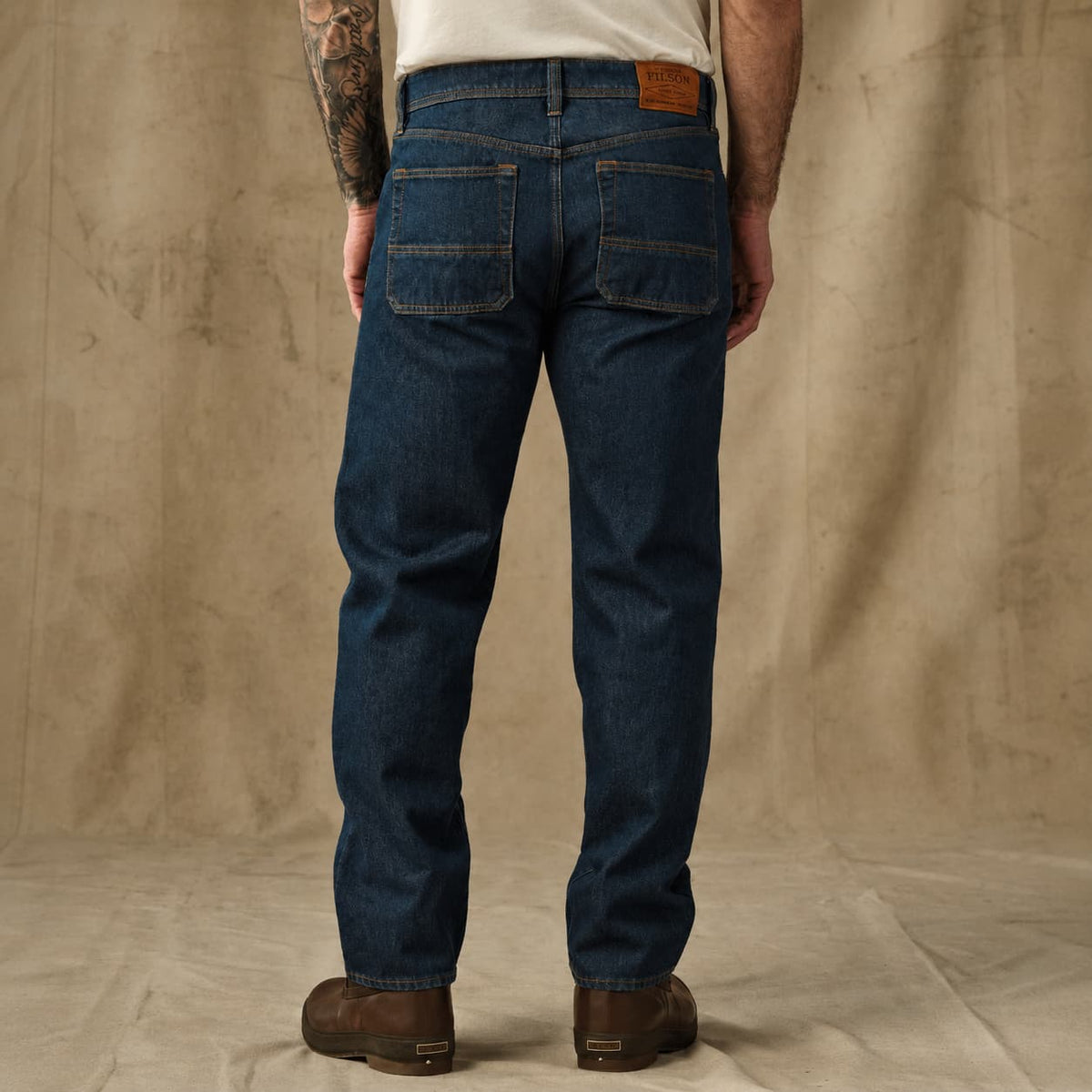 Rear view of a man wearing dark blue Filson denim jeans and brown boots, standing against a textured beige backdrop.