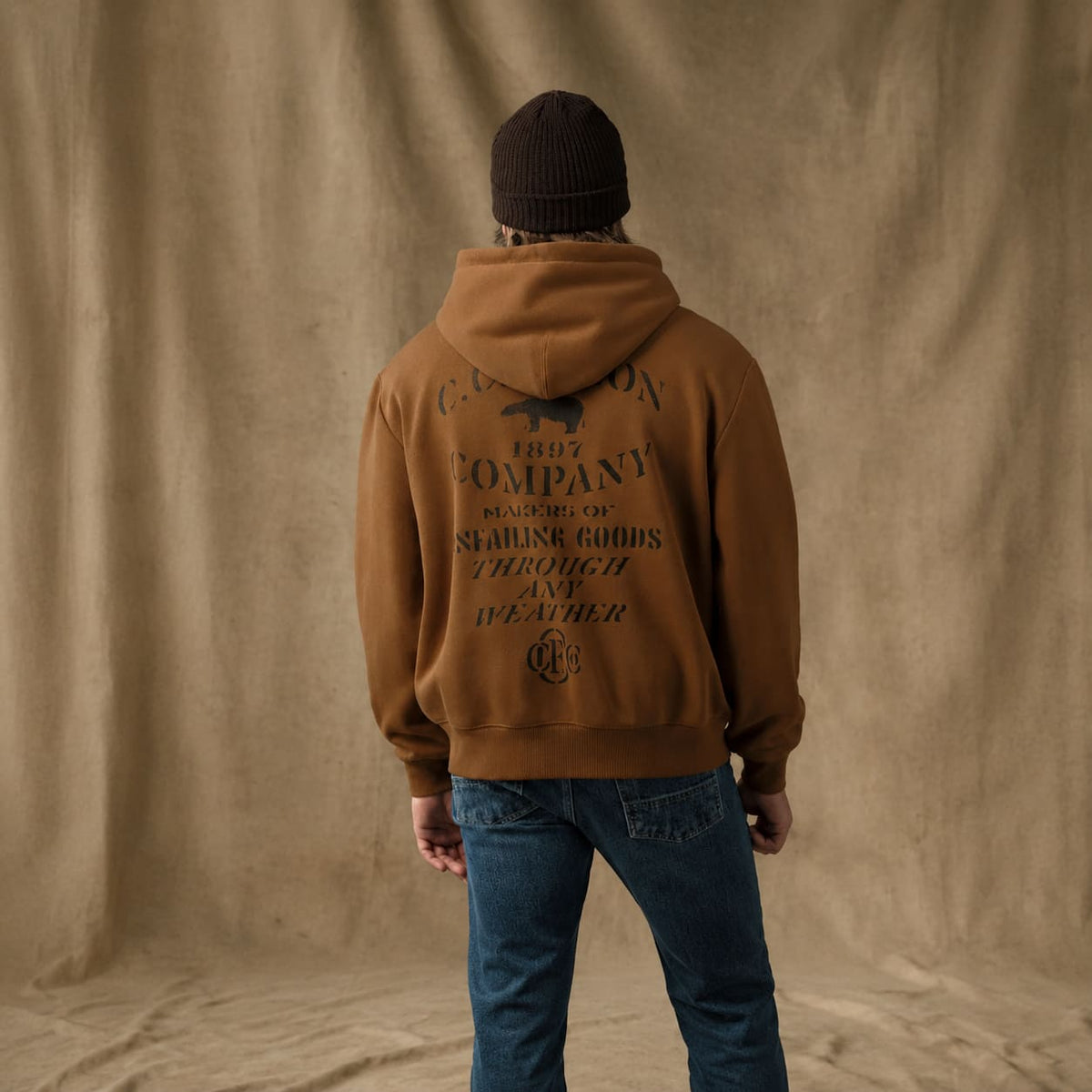 A man viewed from behind wearing a brown Filson hoodie with a large black vintage-style logo and text on the back, paired with blue jeans and a black beanie, standing against a draped tan fabric backdrop.