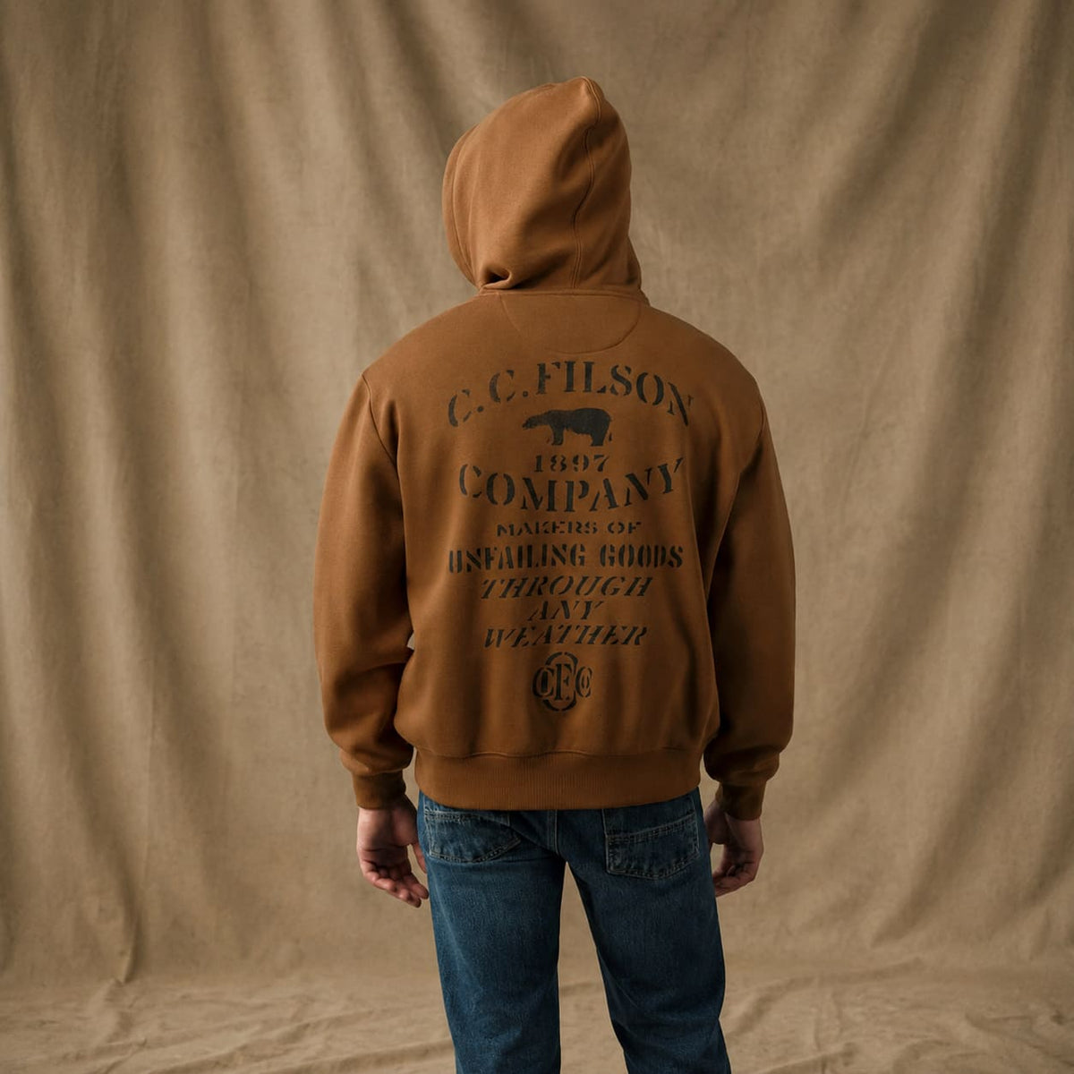 A man viewed from the back wearing a brown Filson hoodie with a large black vintage-style printed logo and text on the back, paired with blue jeans, standing against a draped tan fabric backdrop.