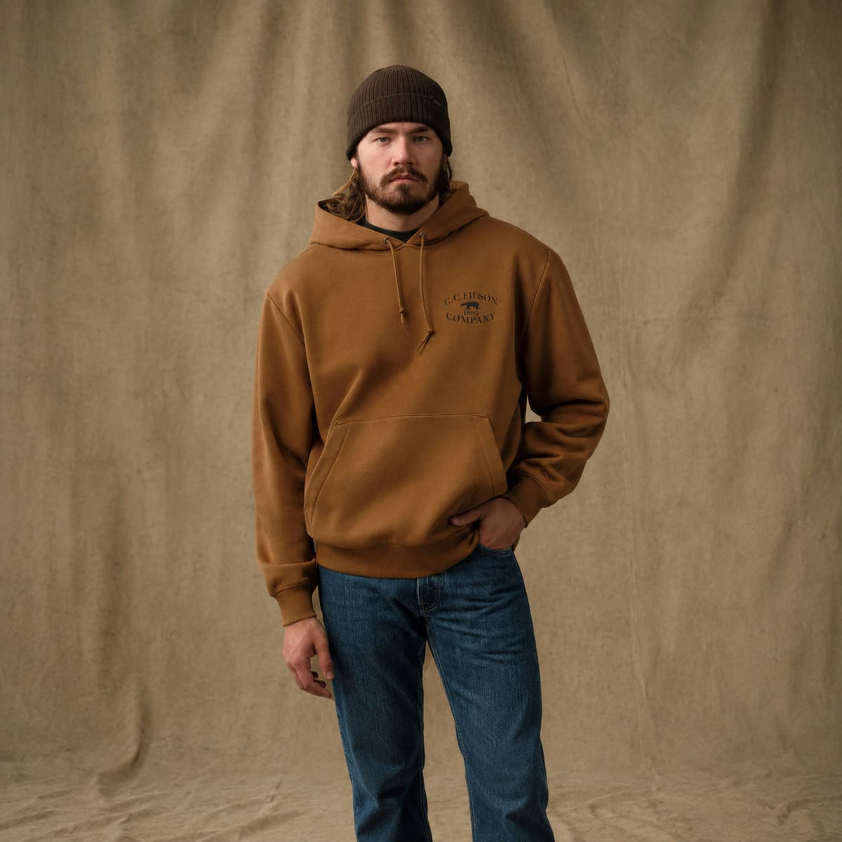 A man wearing a brown ribbed knit beanie, a brown Filson hoodie with a drawstring and kangaroo pocket, and blue jeans, standing against a draped tan fabric backdrop.