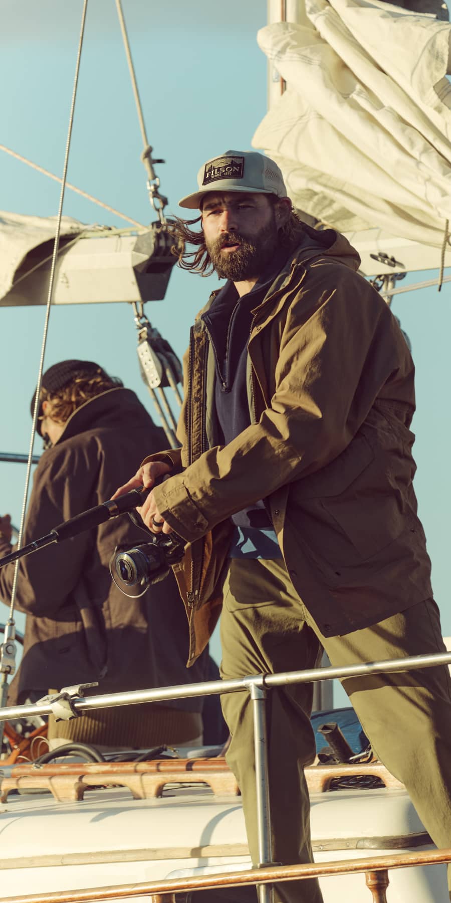 Man on a sailboat holding a fishing rod with another person in the background.