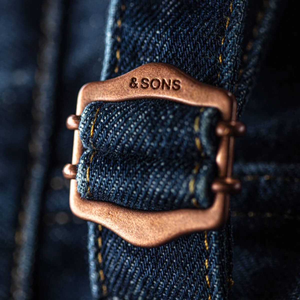 Close-up of a denim strap with a bronze buckle and 'and Sons' engraving.