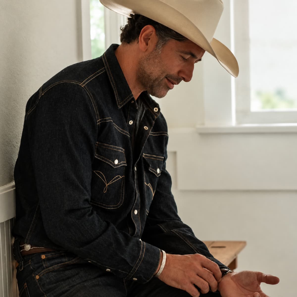A man wearing a dark indigo denim western shirt and matching jeans, sitting indoors next to a window and wearing a cream cowboy hat.