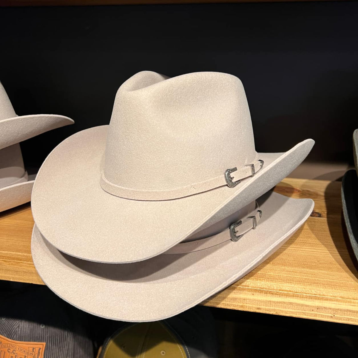 A light beige Stetson felt cowboy hat with silver buckle details, displayed on a wooden shelf next to other hats.