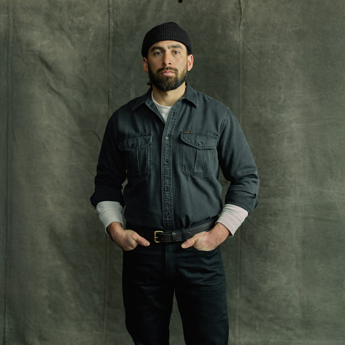 Man wearing a dark denim shirt and black pants against a textured gray background