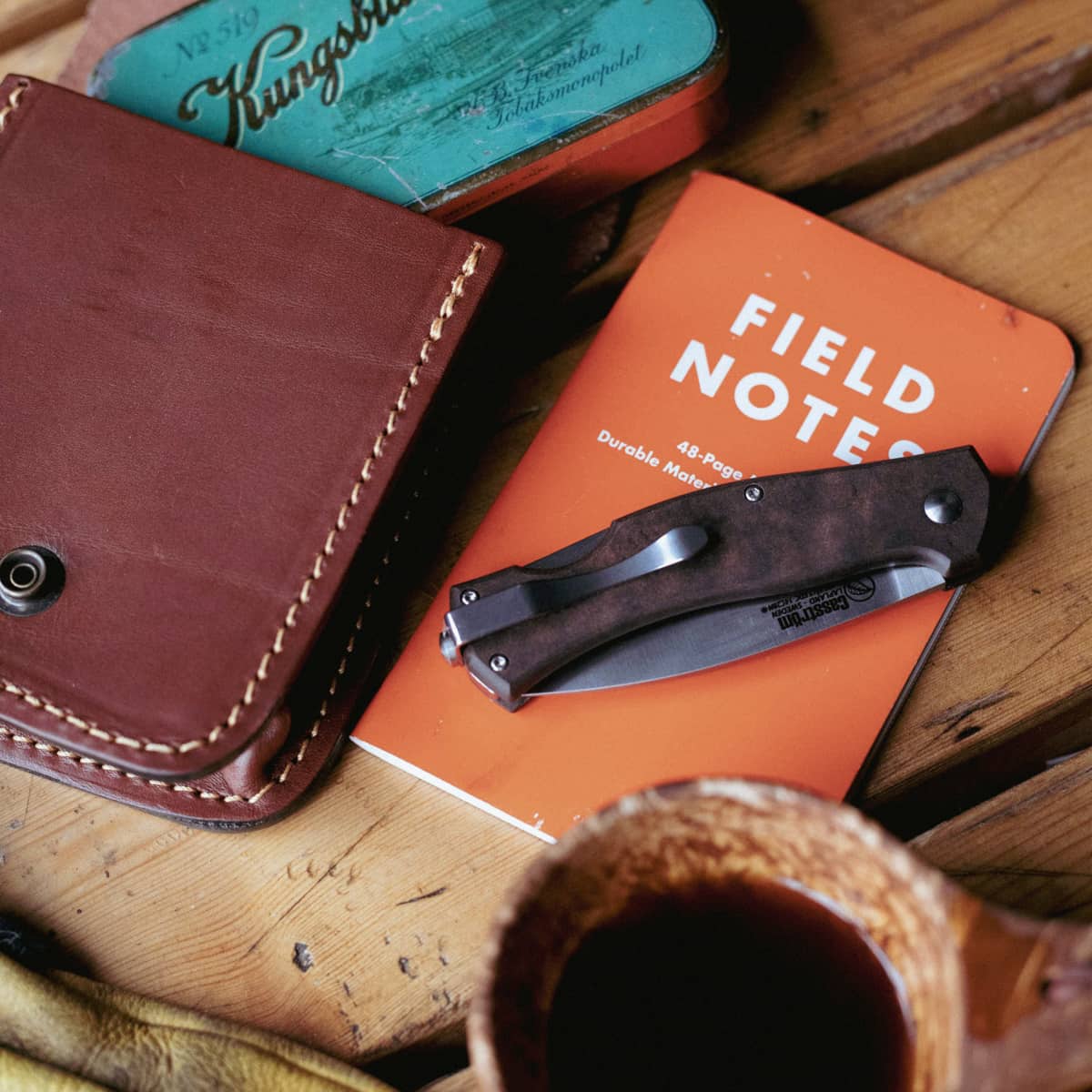 Brown leather wallet, Casstrom knife, and orange Field Notes notebook on a wooden surface.