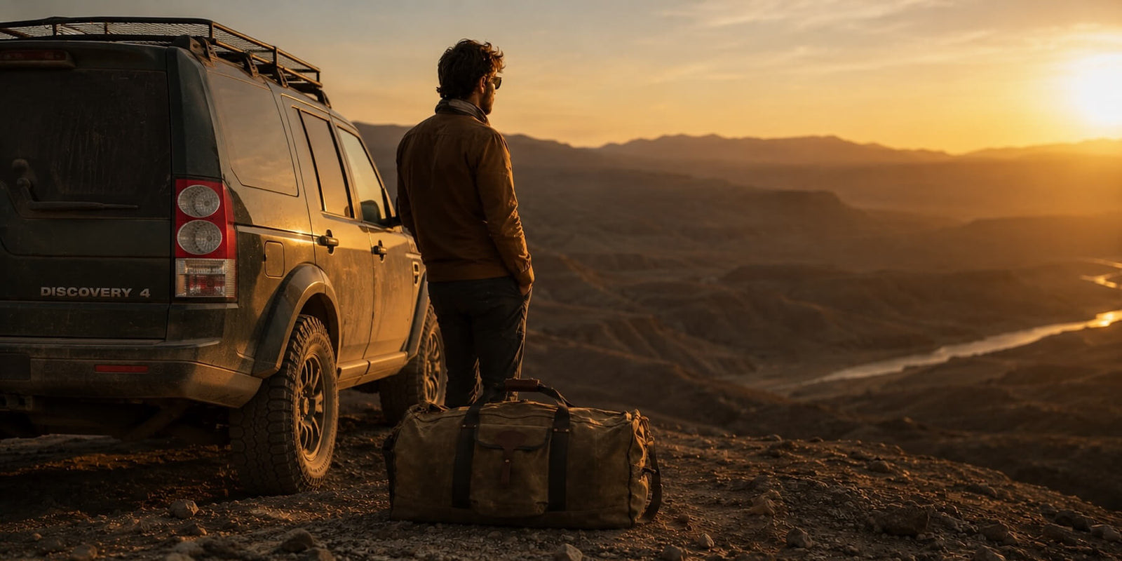 Man standing next to a Land Rover with expedition luggage, overlooking a scenic landscape at sunset.