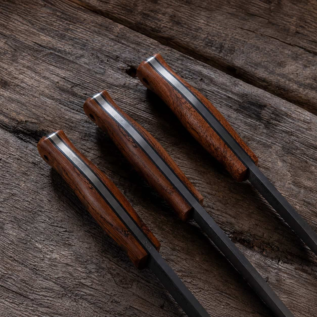 Three bushcraft knives with dark brown wood handles and black blades, arranged diagonally on a rustic dark wood surface.