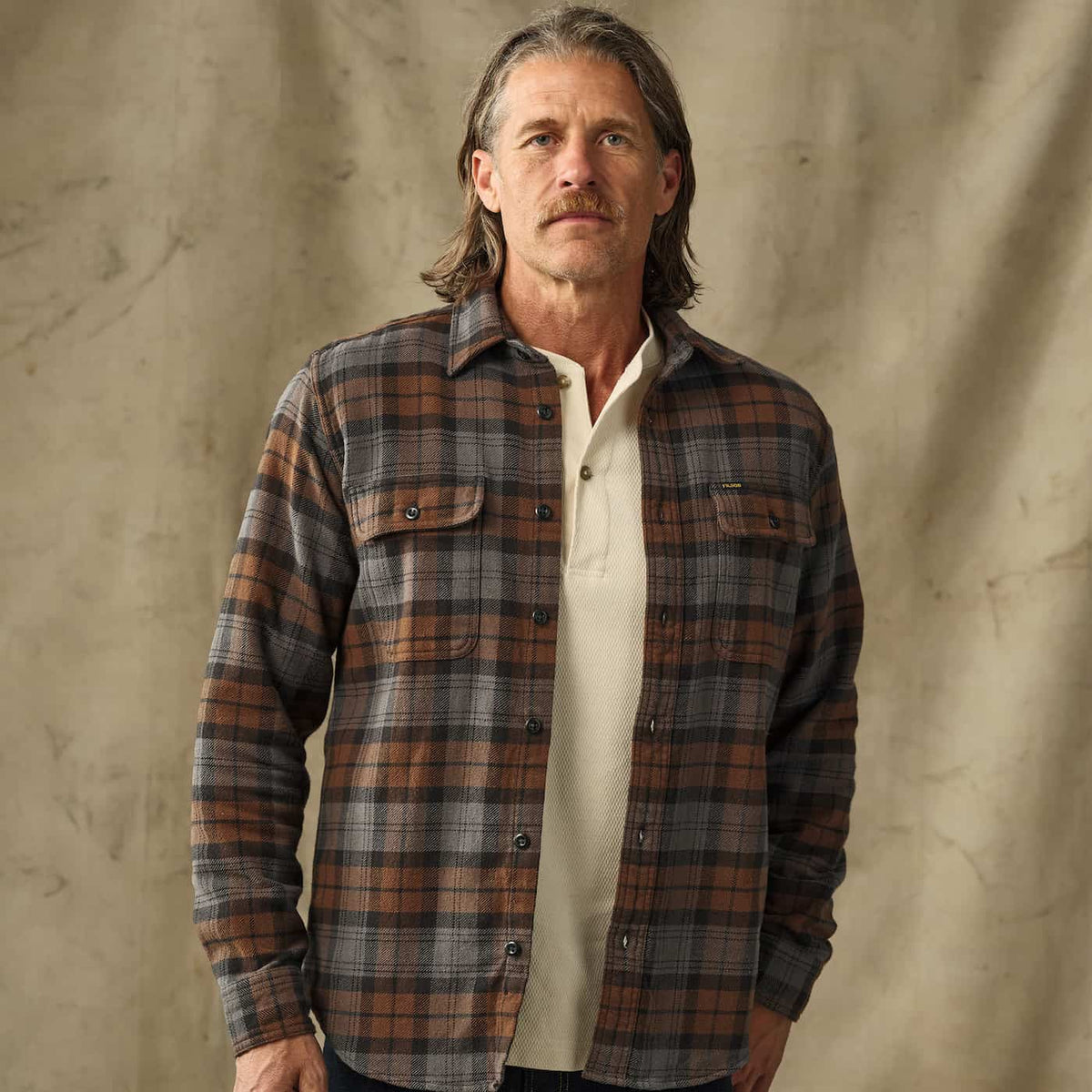 A man wearing a brown and gray plaid Filson flannel shirt over a cream polo shirt, standing against a textured beige backdrop.