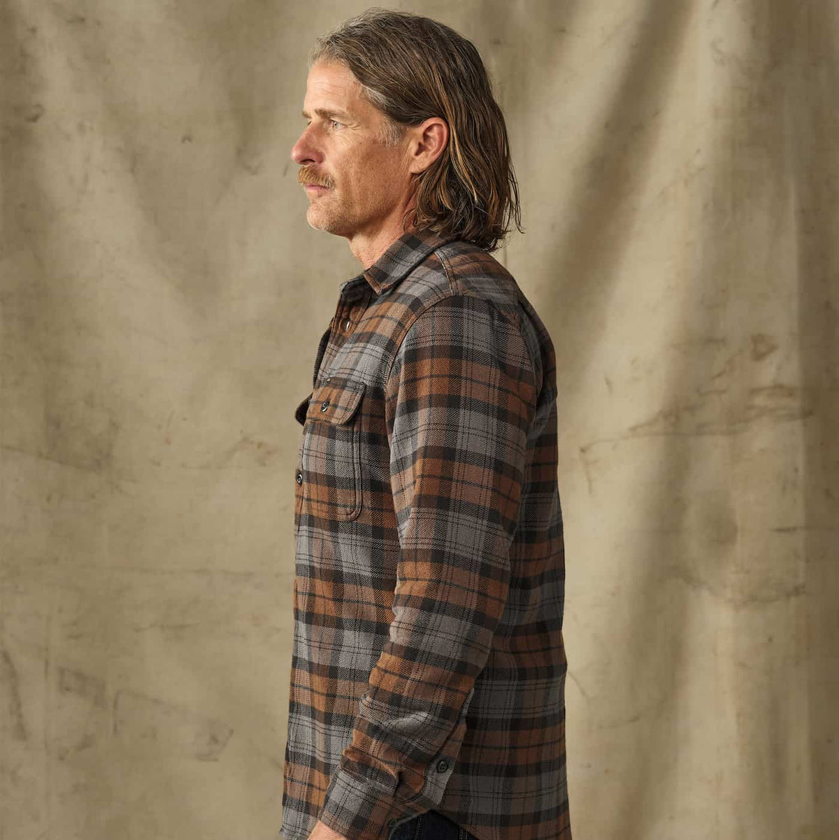 A man in side profile wearing a brown, gray, and black Filson plaid flannel shirt against a textured beige backdrop.