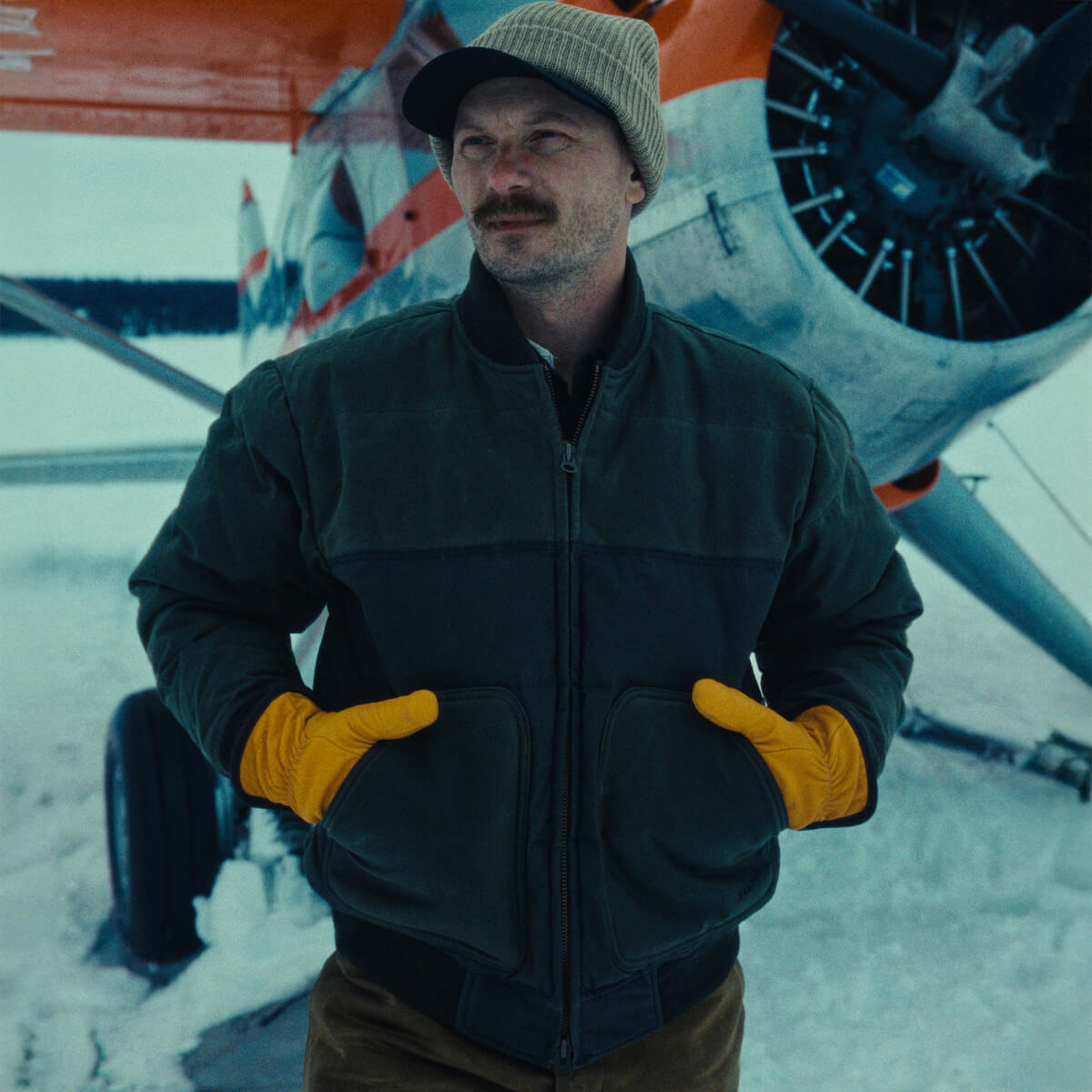 Man wearing a dark jacket, yellow gloves, and a cap standing in front of an airplane in a snowy setting.