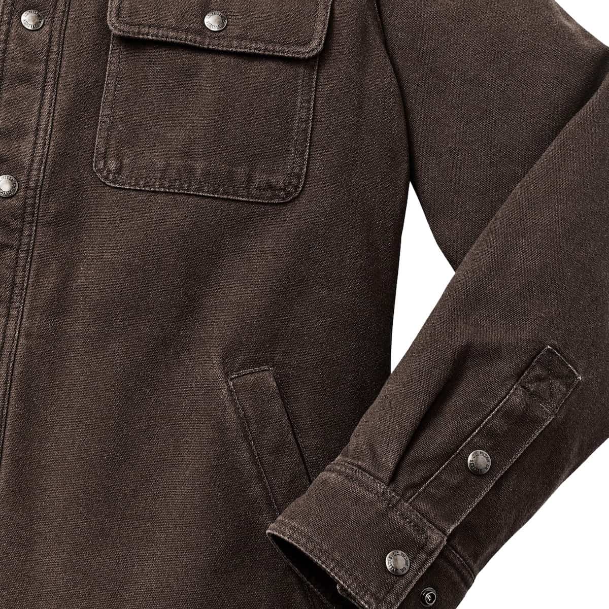 Close-up of a dark brown Filson Fleece Lined Jac-Shirt showing a chest pocket with a silver snap button, the front placket with metal buttons, and the folded cuff of the right sleeve on a white background.