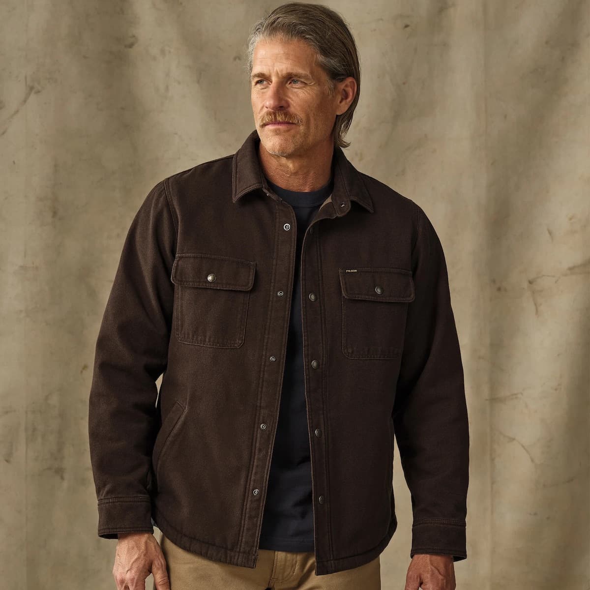 A man wearing a dark brown Filson Fleece Lined Jac-Shirt over a navy blue t-shirt and khaki pants, standing against a textured beige wall.