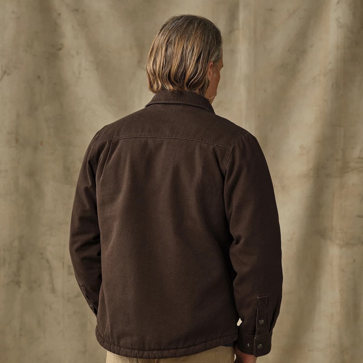 A man viewed from behind wearing a dark brown Filson Fleece Lined Jac-Shirt, standing against a textured beige backdrop.