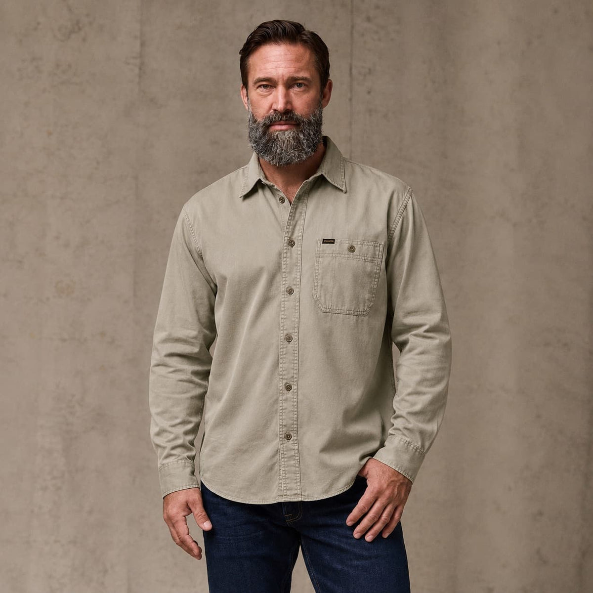A man wearing a light khaki Filson long-sleeve button-down shirt and dark blue jeans, standing against a textured concrete wall.