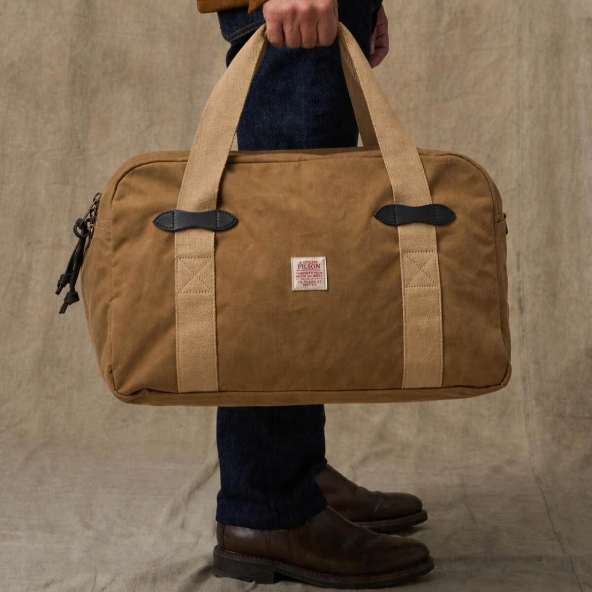 Brown duffel bag with leather straps held by a person against a beige background