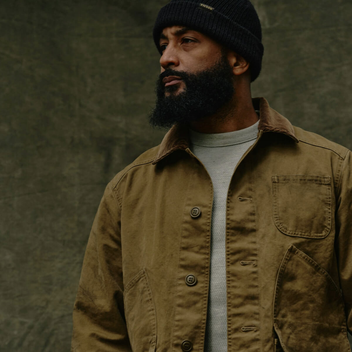 Man wearing a brown jacket and black beanie against a textured green background