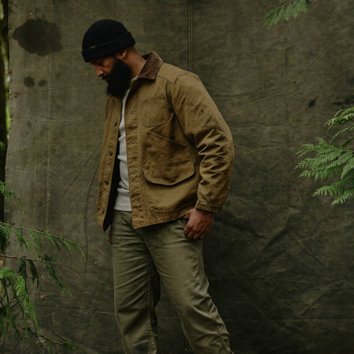 Man wearing a Filson jacket and green pants standing against a textured wall with greenery.
