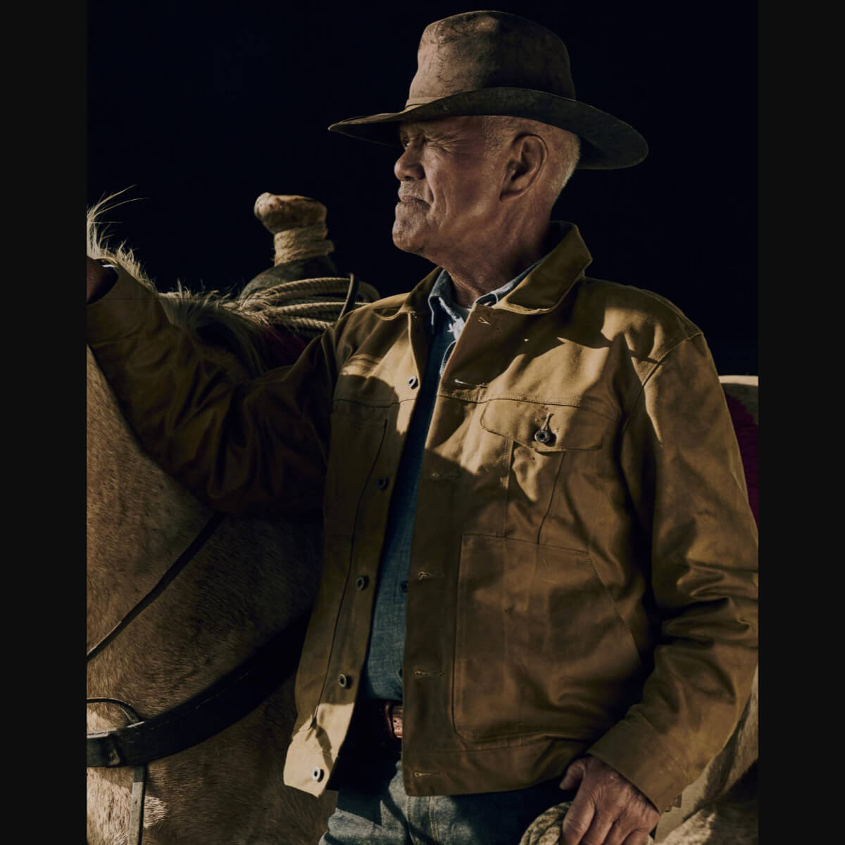 Man in a brown jacket and hat standing next to a horse at night.