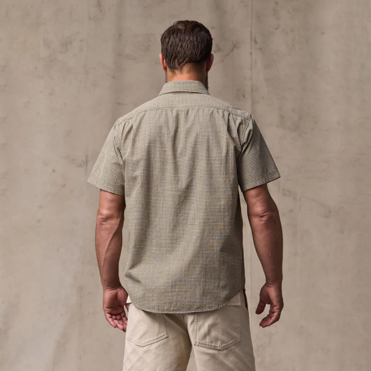 Man wearing a green checkered shirt and beige pants against a beige wall