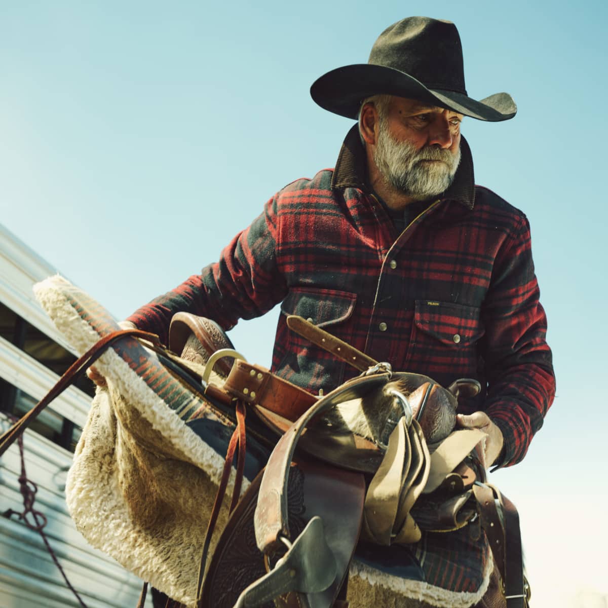 An older man with a white beard wearing a black cowboy hat and a red and black plaid flannel shirt, holding a leather western saddle on a horse against a clear blue sky.