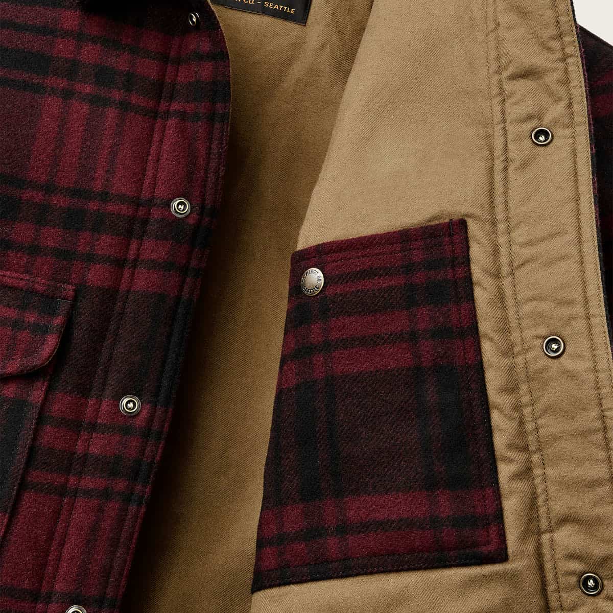 Close-up of a flannel overshirt with a burgundy and black plaid pattern, featuring a snap-button front, metal snap pocket, and tan interior lining.