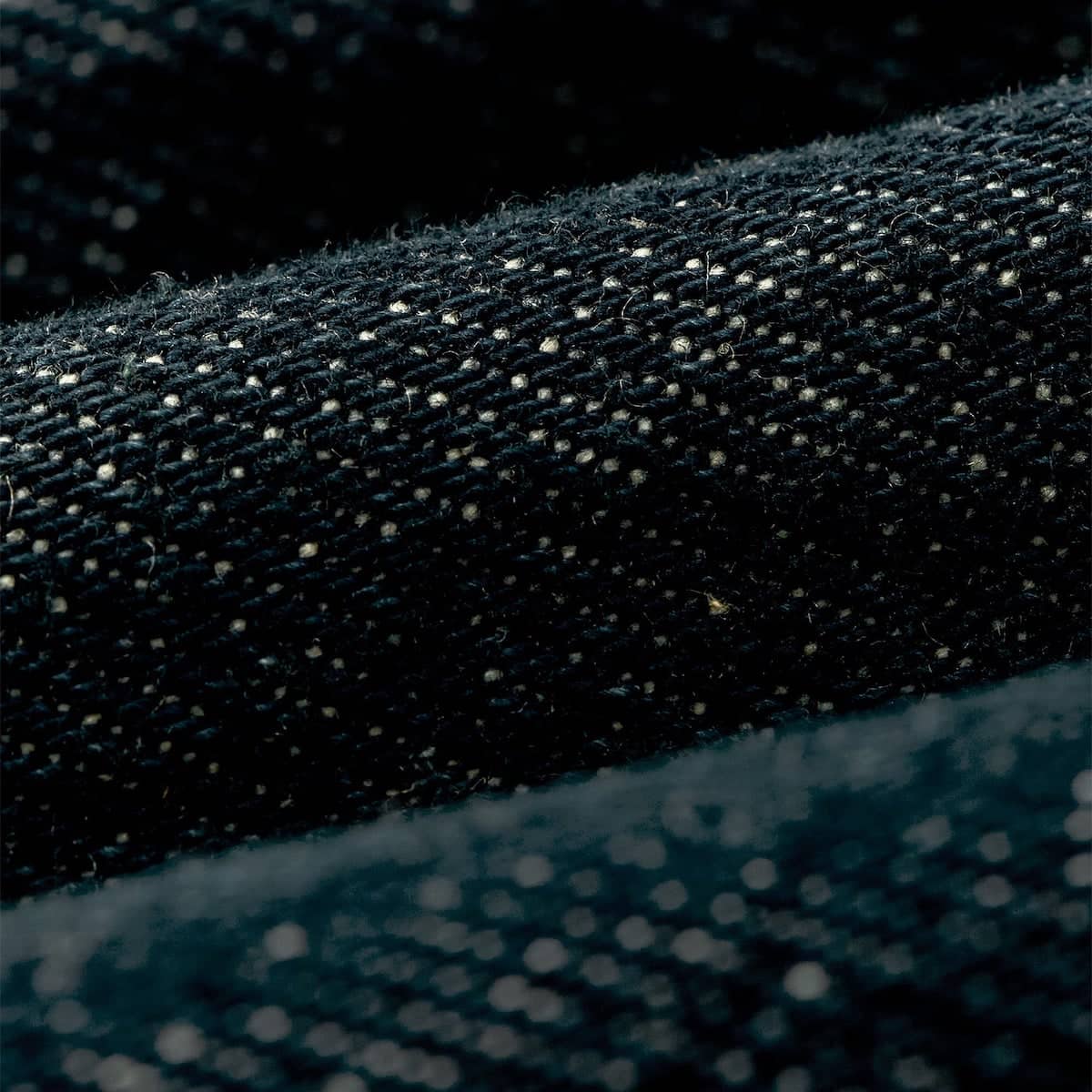 Close-up of dark Filson denim fabric with a woven texture and small light reflective flecks, shown in a rolled material against a dark background.