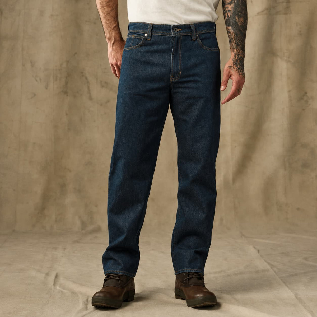 A person wearing dark blue Filson straight-leg denim jeans, a white t-shirt, and brown leather boots, standing against a textured beige backdrop.