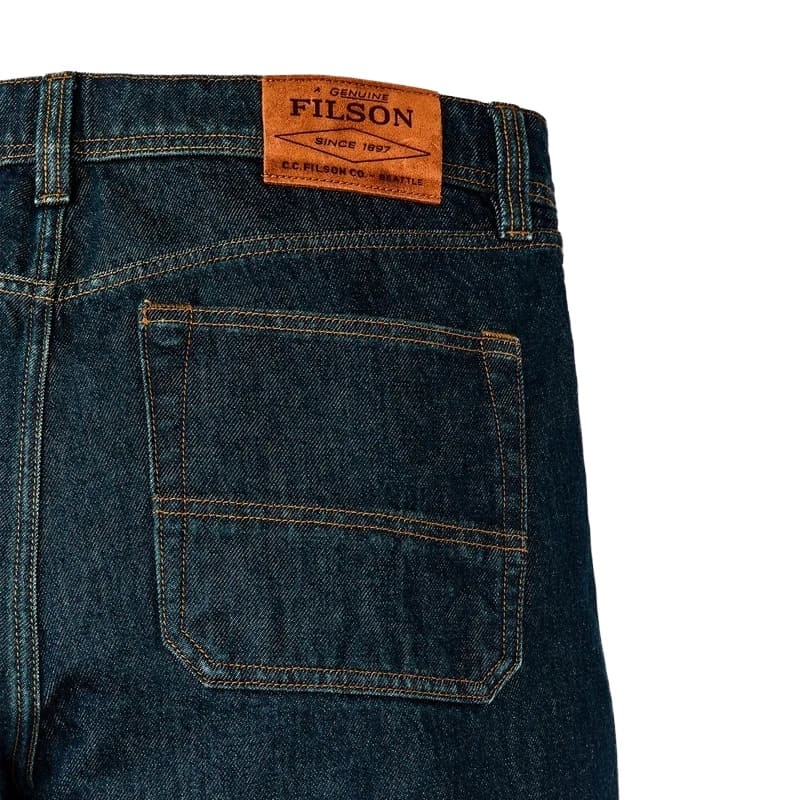 Close-up of the back waistband and pocket of dark indigo denim jeans, showing a tan Filson brand patch and contrast stitching on a white background.