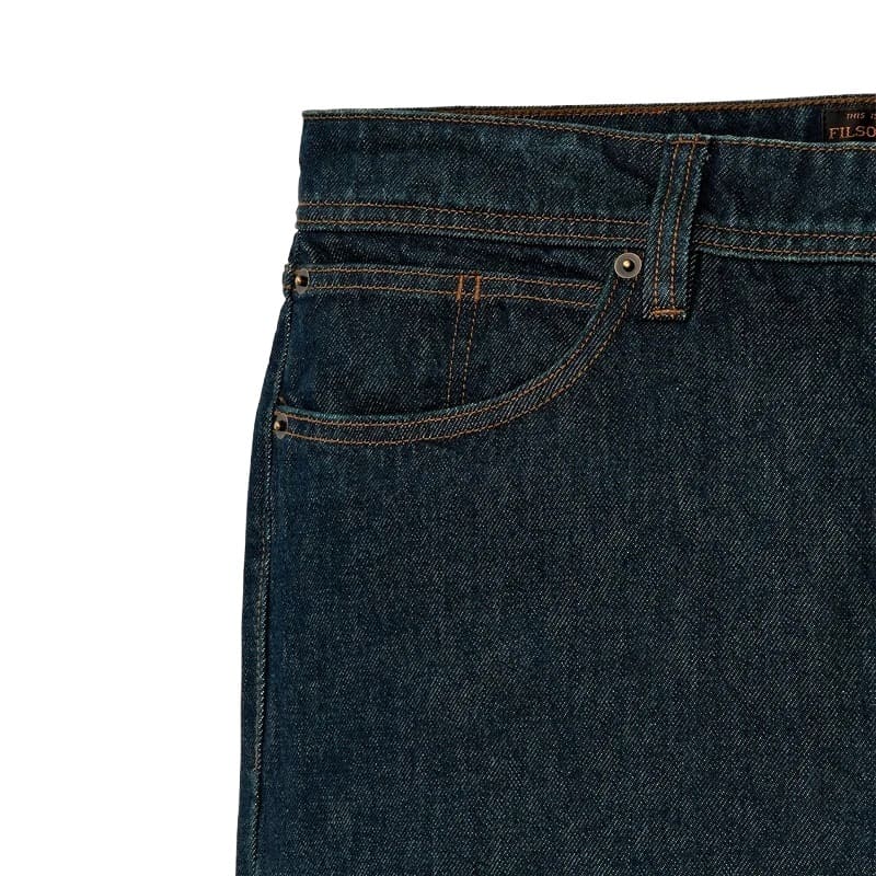 Close-up of the waistband and right front pocket of dark indigo Filson denim jeans, showing contrast tan stitching and a metal button on a white background.