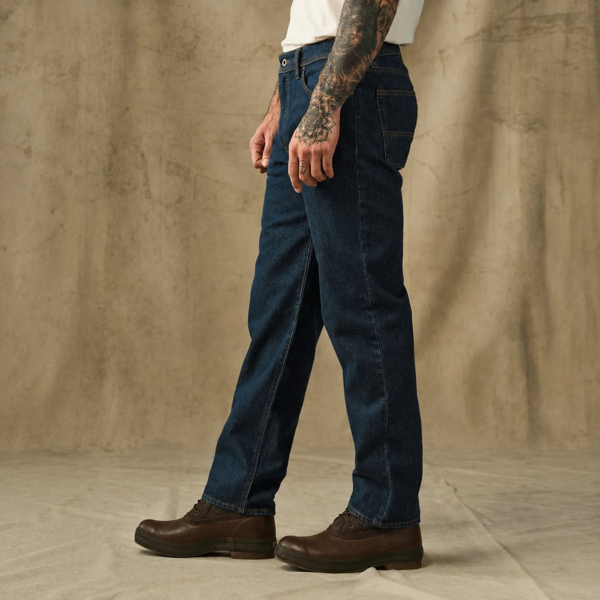 A man wearing dark blue straight-leg Filson denim jeans and brown leather boots, standing against a textured beige backdrop.
