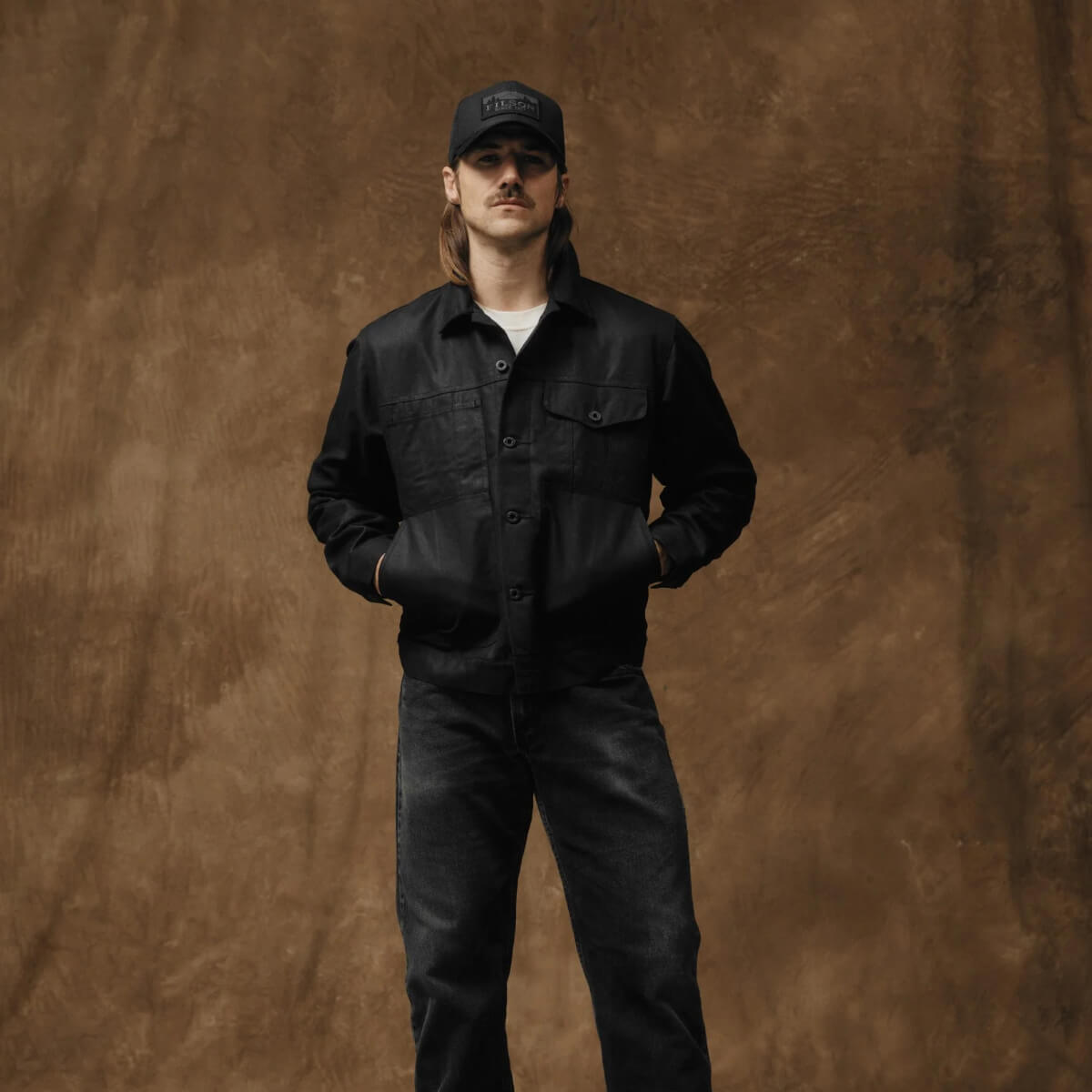 Person wearing a black outfit and cap against a brown background